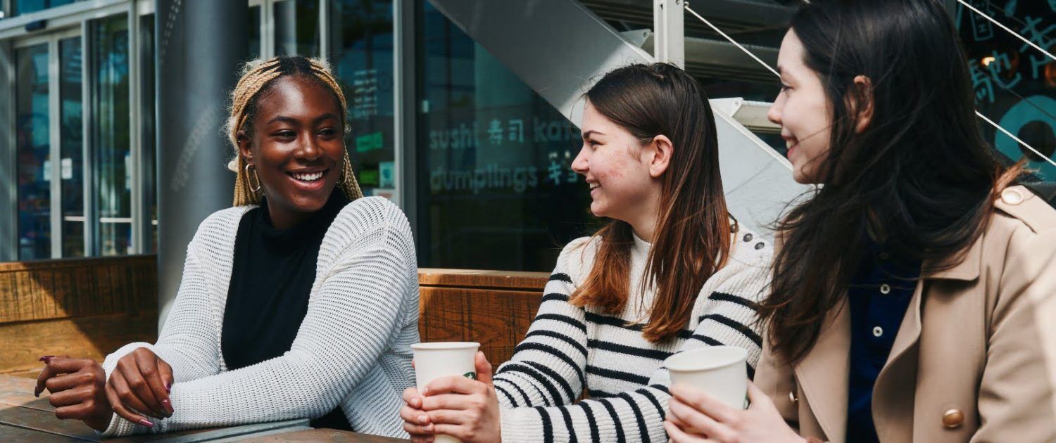 A group of people are sitting around a wooden table on a porch, enjoying a conversation and a meal of sushi, kam dumplings, and other dishes.